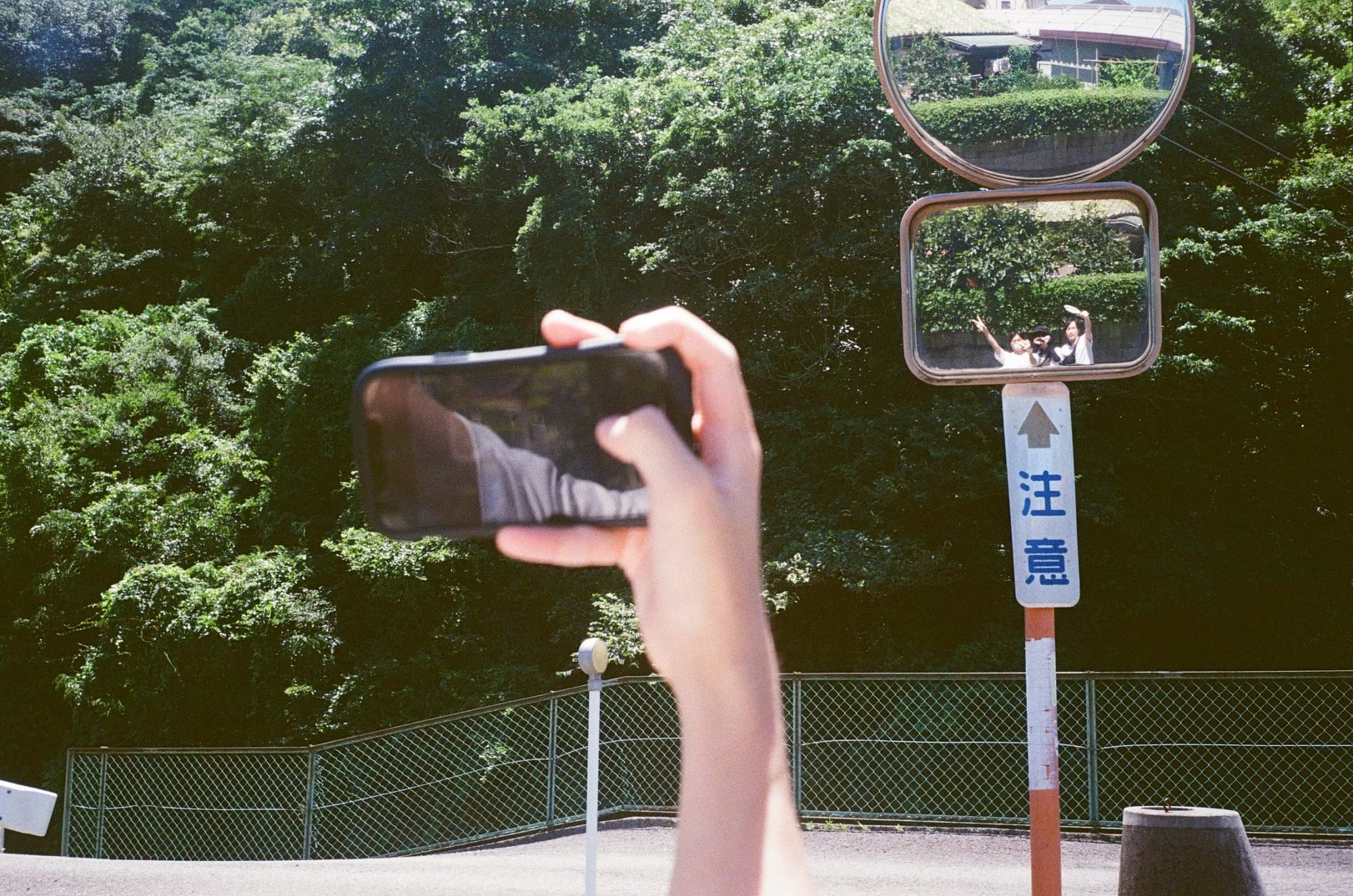 道路の反射鏡に映る、ピースサインをする友人たちをスマートフォンで撮影する様子